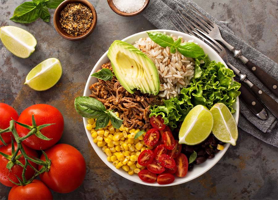 Bol contenant du riz complet, de la viande, de l'avocat, du maïs, des tomates cerises et des haricots noirs, illustrant les glucides du riz complet.