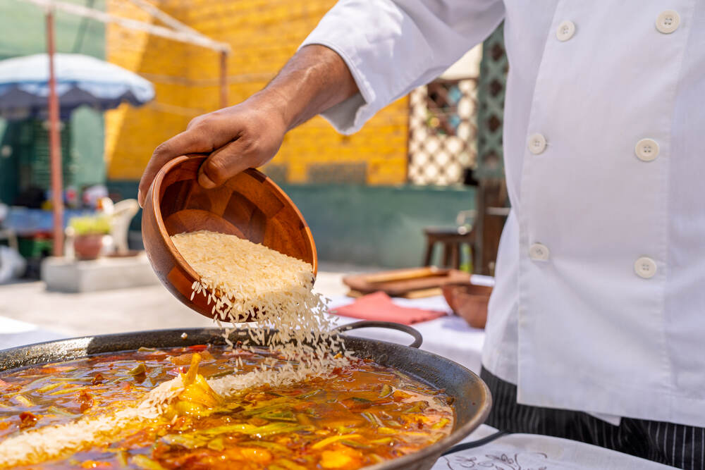 طاهٍ يسكب الأرز النيئ في مقلاة باييلا كبيرة في الهواء الطلق، موضحًا مرحلة أساسية في طهي الأرز للباييلا