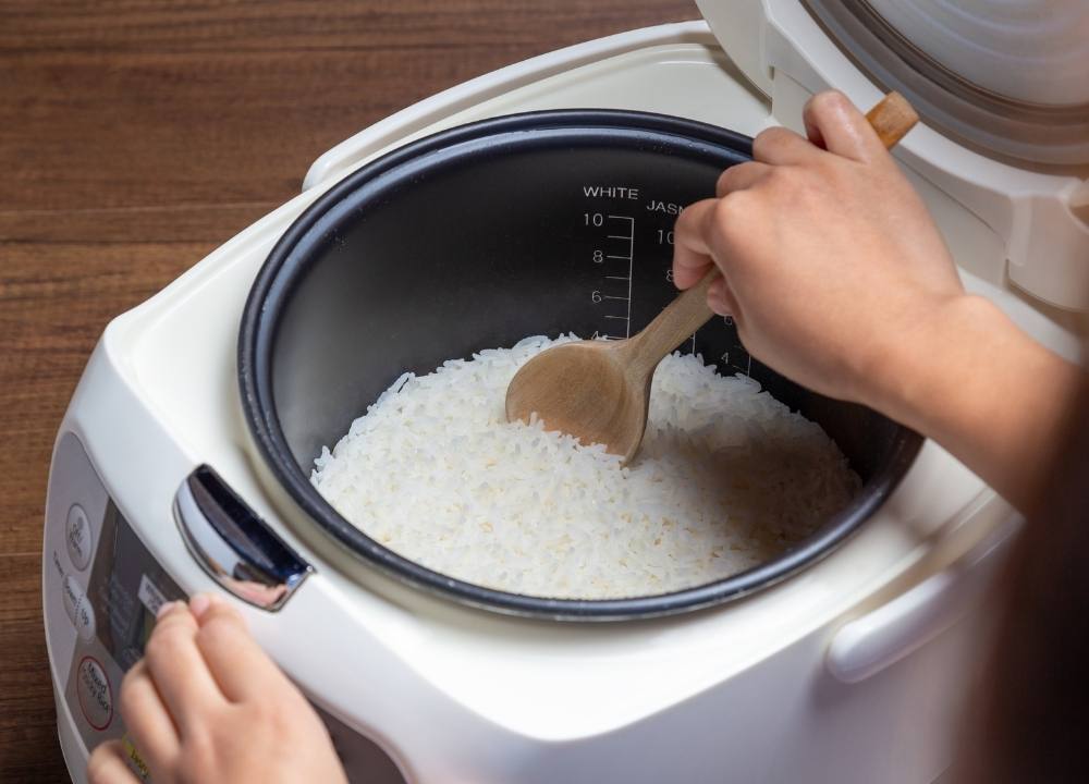شخص يحرك الأرز الأبيض المطهو في جهاز طهي الأرز بملعقة خشبية. القيم الغذائية للأرز