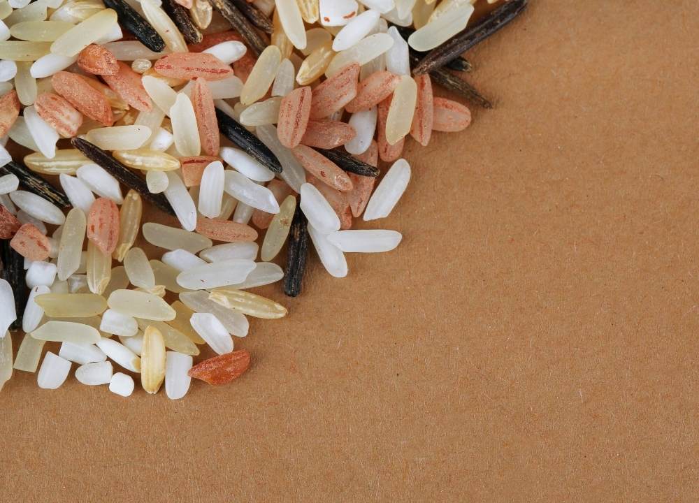 صورة مقربة لمزيج من حبوب الأرز النيء، بما في ذلك الأرز الأبيض والبني والأسود (أرز بري)، مبعثرة على سطح بني.