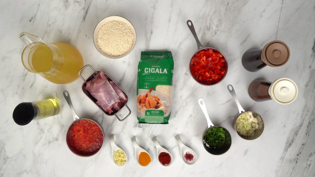 Tous les ingrédients sont disposés sur une table pour préparer une paella au lapin : riz, viande de lapin, légumes coupés, épices et bouillon.