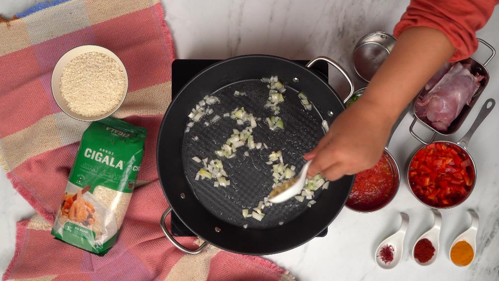 Dans une grande poêle noire, des oignons hachés sont en train d'être sautés dans de l'huile, première étape de la préparation de la paella.