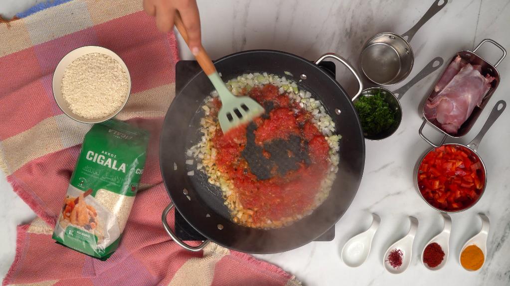 Une spatule mélange une sauce tomate épaisse avec des oignons hachés dans une poêle, formant la base aromatique de la paella.