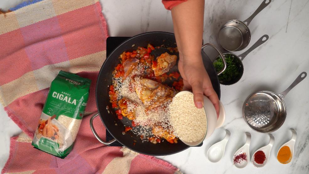 Du riz est ajouté à la poêle contenant des morceaux de lapin et des légumes déjà en train de cuire pour la paella.