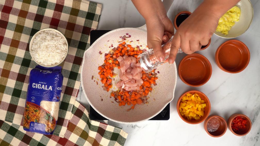 Ajout de morceaux de poulet et de carottes dans une poêle pour la préparation du Nasi goreng, à côté d'un paquet de riz Cigala.