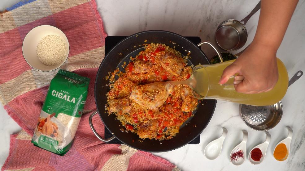 Une personne verse délicatement du bouillon de poulet dans une grande poêle contenant du riz, du lapin et des légumes pour la paella.