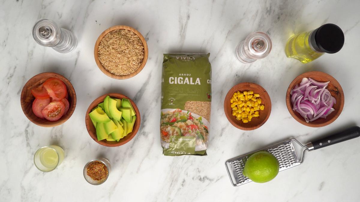 Ingrédients pour la salade d'avocat et de tomate : riz complet Cigala, avocat, tomate, maïs, oignon rouge, et citron vert disposés sur une table en marbre.