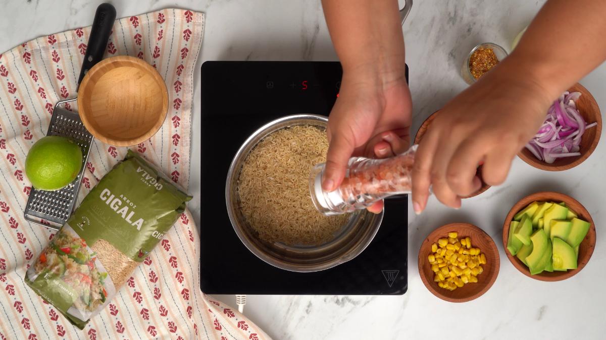 Cuisson du riz complet Cigala pour la salade. Une main ajoute du sel dans la casserole sur une plaque à induction.