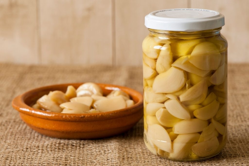 مرطبان كبير يحتوي على فصوص ثوم محفوظة في الزيت، موضوع بجانب وعاء صغير من الفخار.