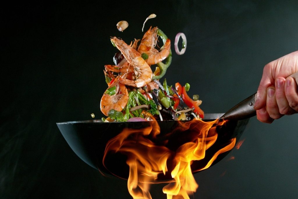 لقطة مقرّبة لوك/مقلاة تتصاعد منها ألسنة لهب مع روبيان وخضار تُقلى في الهواء الطلق.