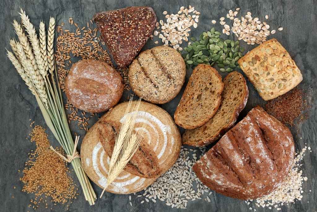 فوائد النظام الغذائي الماكروبيوتيكي منظر علوي لتشكيلة من الخبز الكامل والحبوب (القمح، الشوفان والبذور) موضوعة على سطح داكن.