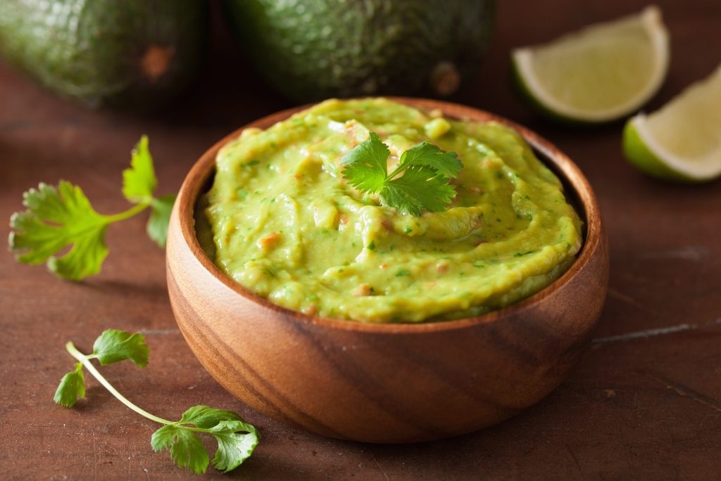 Guacamole et chips Gros plan sur un bol en bois de guacamole, garni de coriandre, avec des avocats et des citrons verts en arrière-plan.