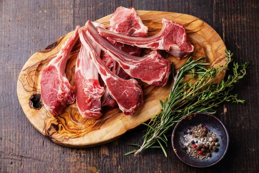 ضلوع لحم غنم نيئة موضوعة على لوح تقطيع خشبي مع إكليل الجبل وتوابل متنوعة.