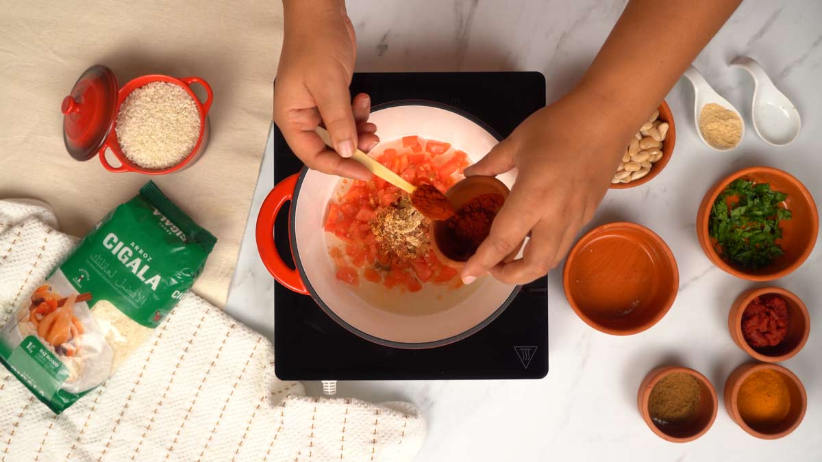 Préparation de la base aromatique Une personne ajoute des épices en poudre aux tomates qui cuisent dans une casserole.