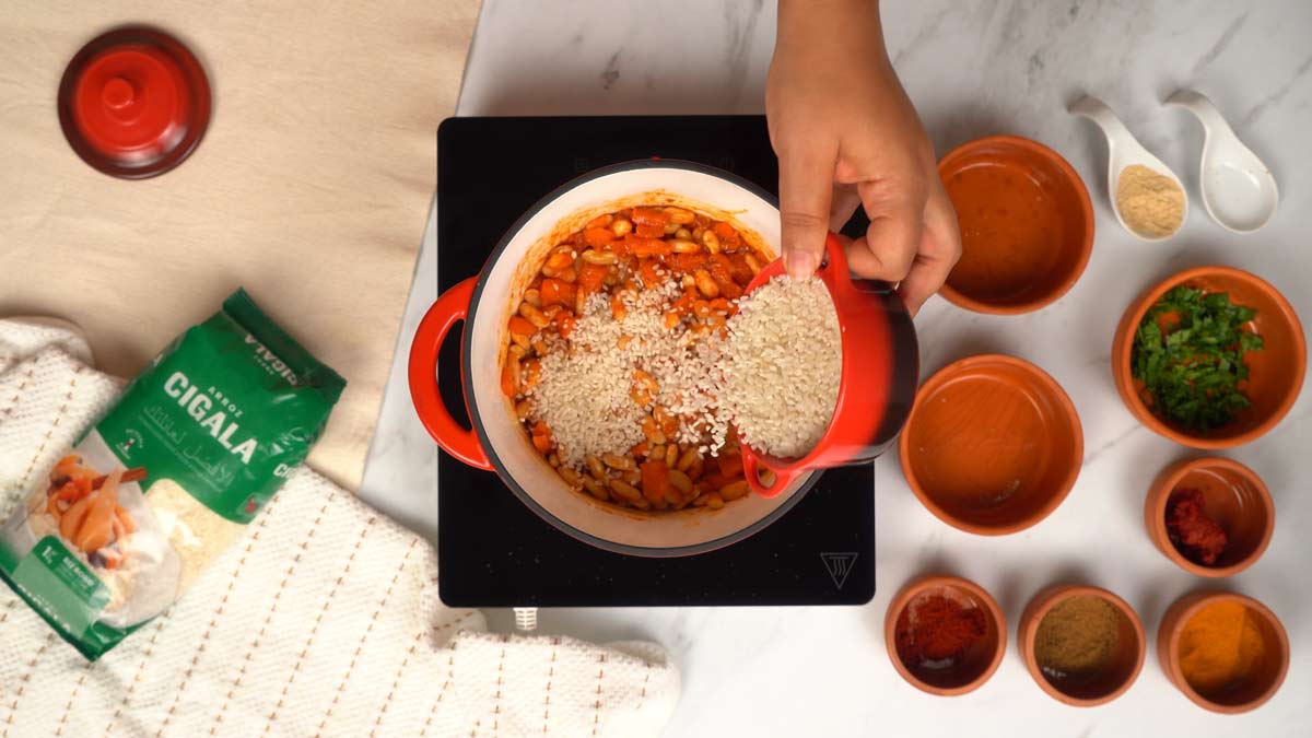 Ajout des haricots blancs et du riz Une personne verse un bol de riz cru sur la sauce et les haricots blancs dans la casserole.