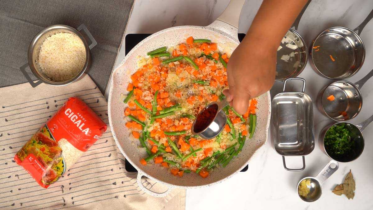 إضافة الأرز والتوابل شخص يضيف البابريكا والتوابل إلى الأرز والخضروات في المقلاة.