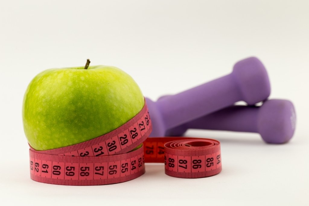 التحكّم في الوزن تفاحة خضراء يحيط بها شريط قياس وردي، مع أثقال رياضية بنفسجية غير واضحة في الخلفية.