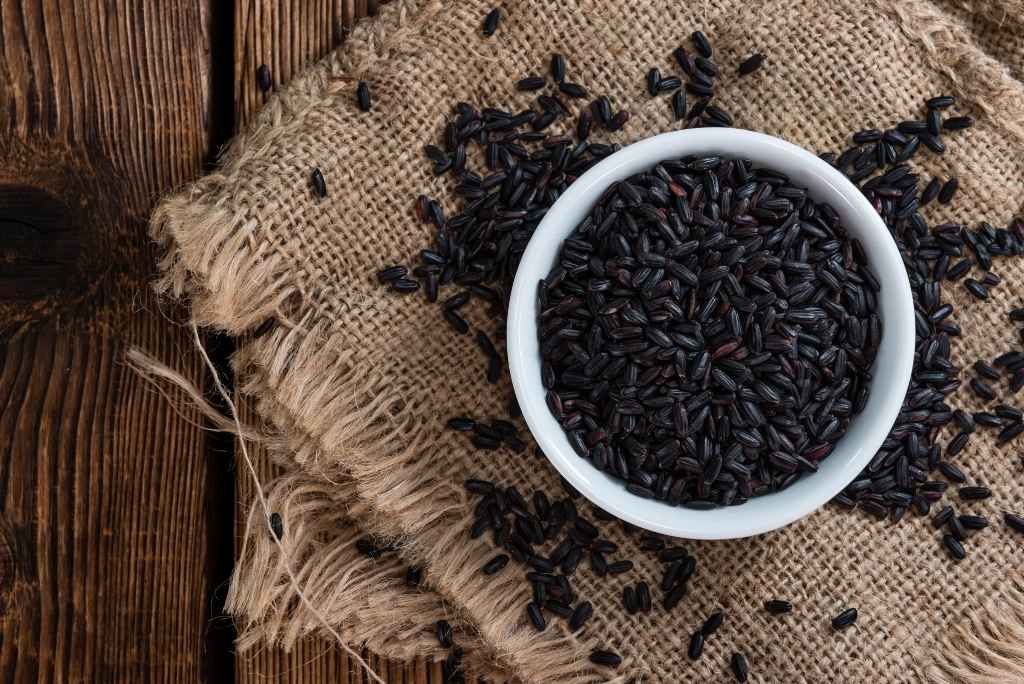 ما هو الأرز الأسود ولماذا يُعتبر "ممنوعاً"؟ منظر علوي لوعاء يحتوي على أرز أسود نيّئ فوق قماش من الخيش وعلى طاولة خشبية.