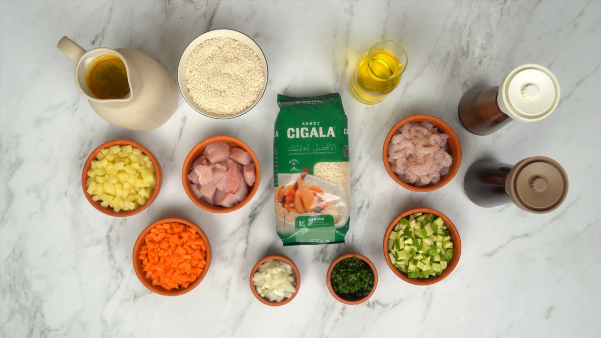 Préparation des ingrédients On prépare l'ensemble des ingrédients, notamment le riz rond, les crevettes, les dés de poulet, ainsi que divers légumes coupés comme les carottes, les courgettes et les pommes de terre.