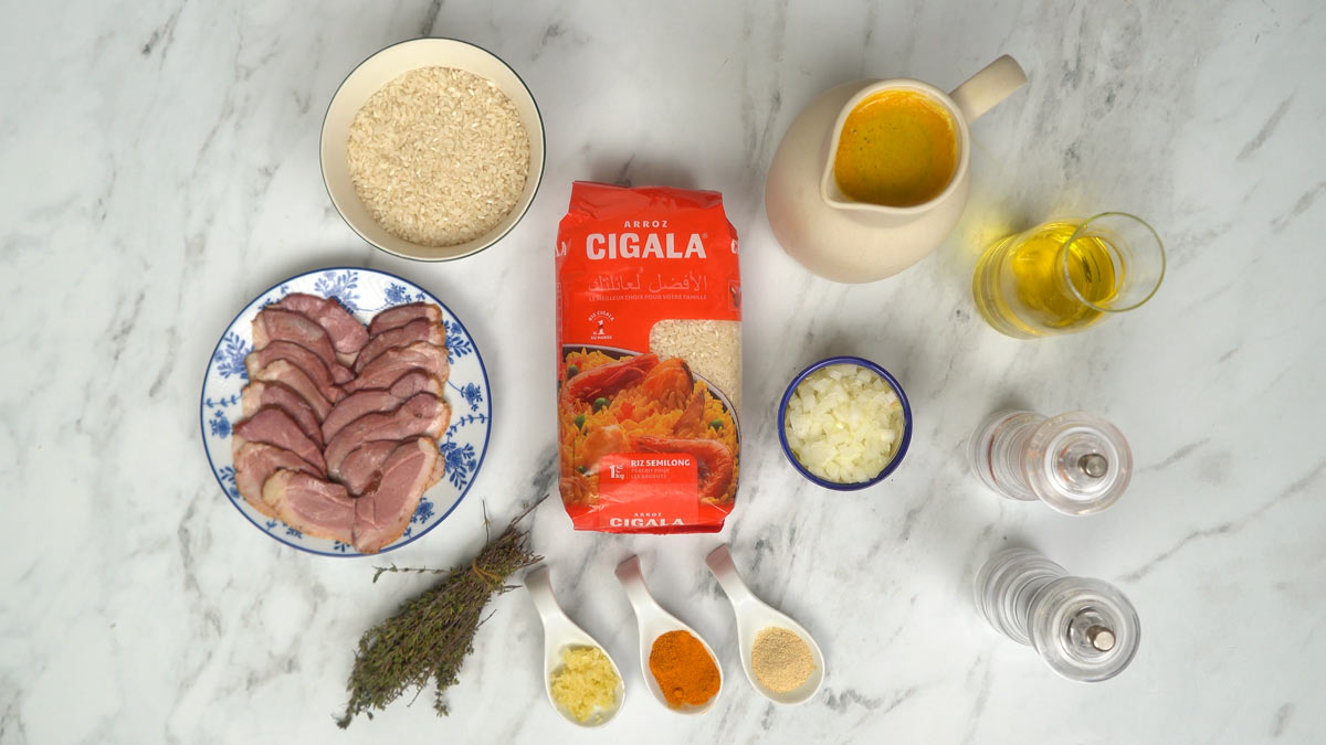 On prépare les ingrédients, notamment des tranches de magret de canard fumé, du riz, du bouillon de volaille, de l'oignon haché et des épices.