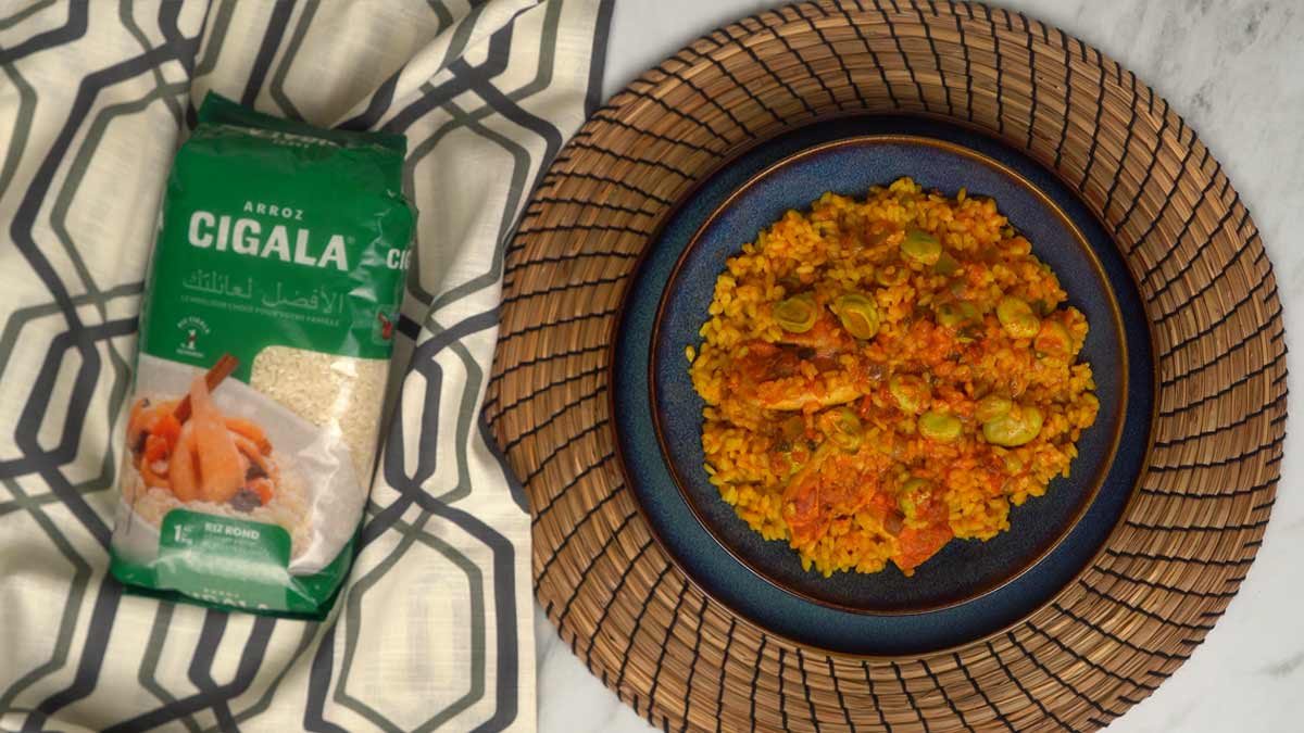 Le plat terminé offre un riz onctueux et coloré, où les morceaux de lapin sont servis avec les fèves, le tout présenté sur une assiette bleue foncée.